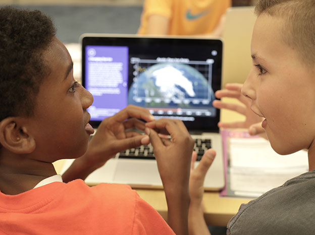 Two elementary students discussing in front of a computer with an image of the earth