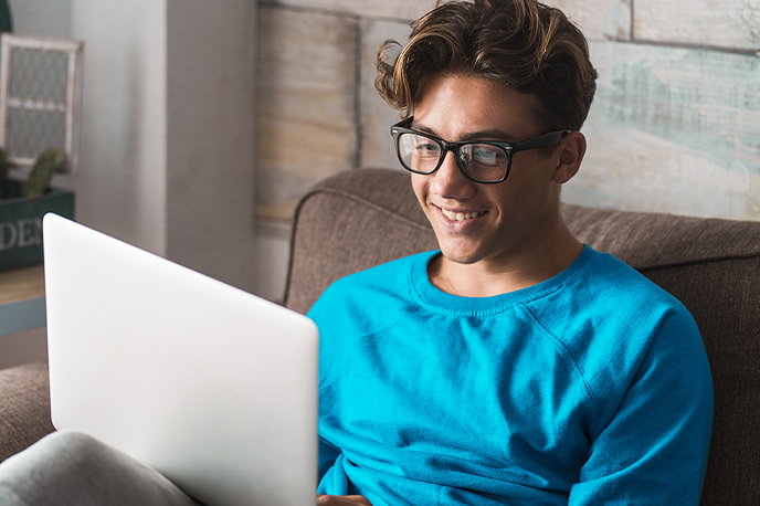 A high school student smiling and working on a laptop while sitting on a couch