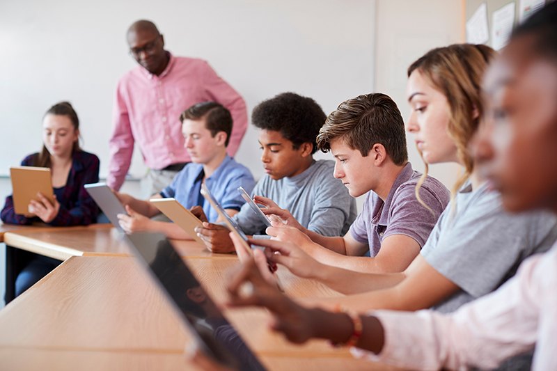 Group of high school students working on tablets while teacher oversees in the background