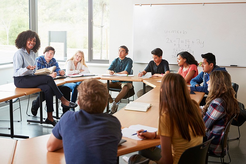 Group of high school students seated at their desks and surrounding an enthusiastic teacher