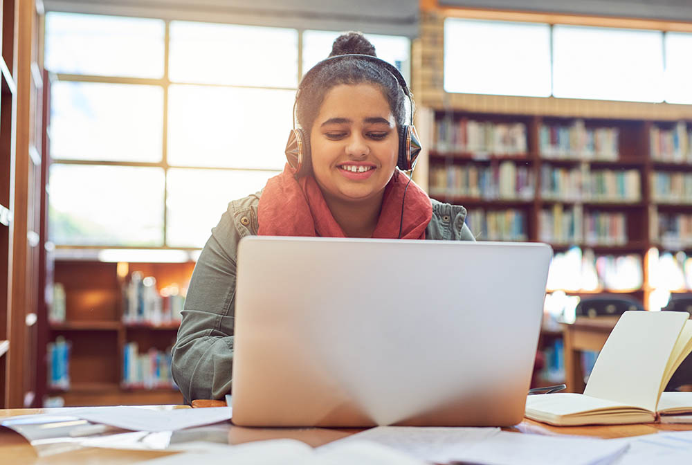 Shot of a cheerful young female student working on a laptop while listening to music with her headphones inside of a library