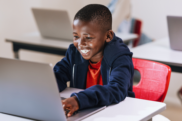 Elementary student smiling and typing on laptop in a classroom.