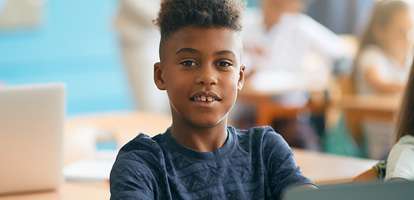 Elementary school student seated at a desk in a classroom while working on a laptop and smiling at camera