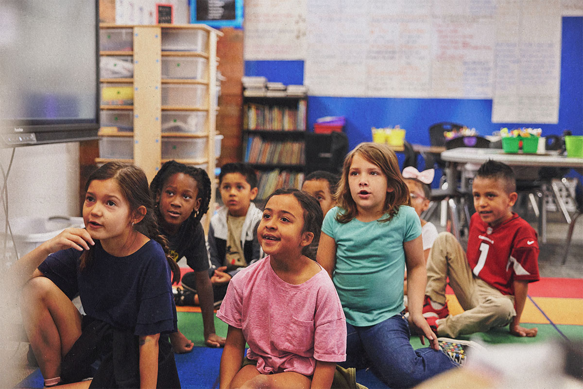 A group of elementary students sits on a colorful classroom rug, looking toward the front of the room.