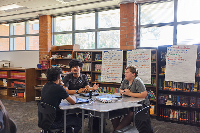 In a light-filled room, a group of three middle school boys sit at a table and talk.
