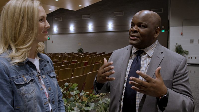A woman, Lauren, talks to a man, Dr. Dawson, in an empty auditorium.