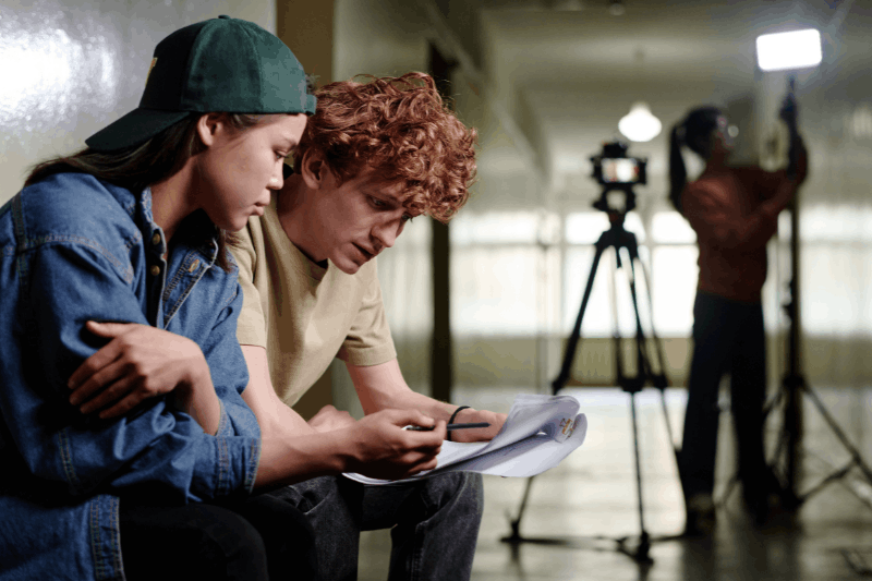 Two students look at a script with camera and lighting equipment in the background