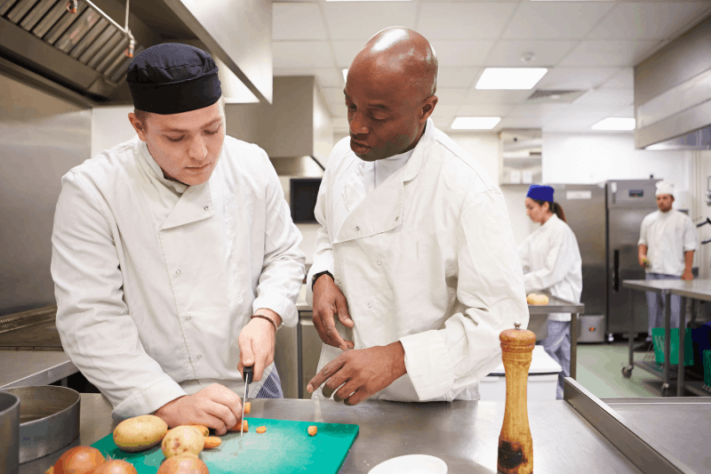A chef is teaching his student how to chop carrots 