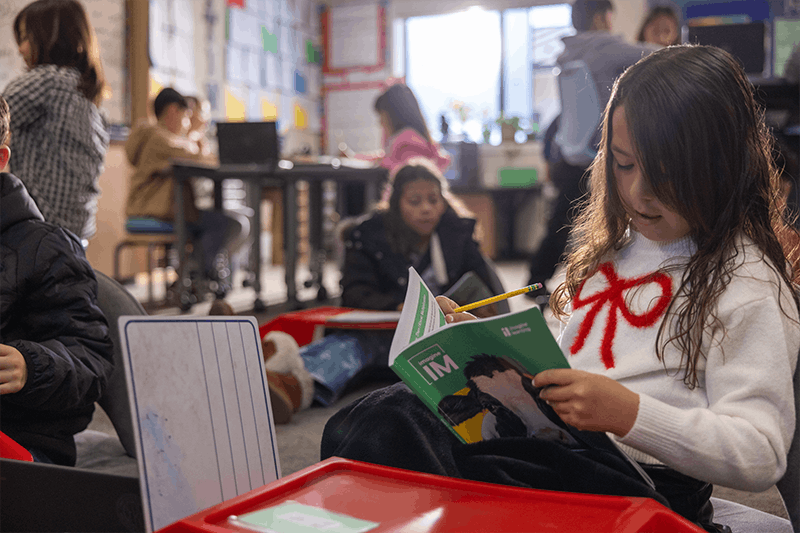 a young student working in a workbook while students are working in groups in the background
