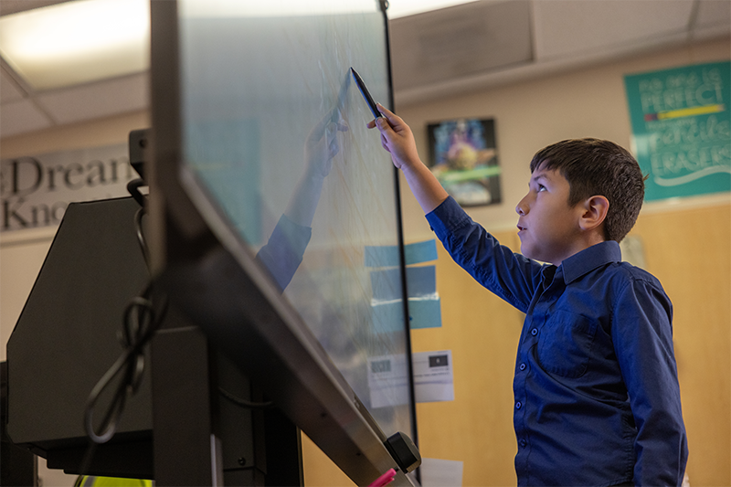 a young student stands in front of a classroom board, writing on the board with a stylus